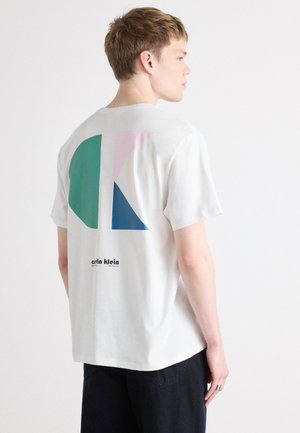 Bela bombažna majica s geometrijskim vzorcem na hrbtu, ki vključuje modre, zelene in rožnate oblike, ter napis "calvin klein" natisnjen spodaj v črni barvi.