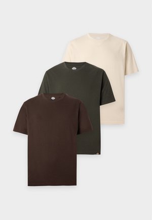 Junger Mann steht mit Händen in den Taschen, trägt ein dunkelbraunes T-Shirt, schwarze Hose, schwarz-weiße Sneakers und einen schwarzen Umhängegurt.