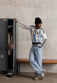 Uma pessoa vestindo uma camisola desportiva às riscas e jeans inclina-se sobre um armário aberto com uma bola de basquete em uma rede pendurada dentro, ao lado de um banco de madeira.