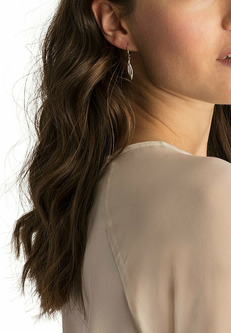 Silverörhängen med droppform och bladdesign, med fina detaljer och en polerad yta, bärs av en modell med vågigt brunt hår.