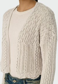 Cardigan lavorato a maglia beige chiaro con apertura frontale. Presenta un motivo in pizzo e orlo ondulato. Indossato sopra una maglietta chiara e jeans di denim.