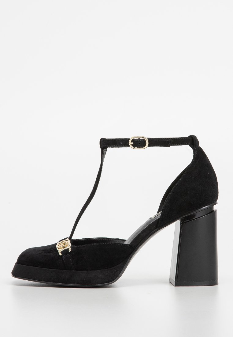 Zapato de tacón alto de ante negro con un diseño de tira en T y una hebilla decorativa dorada. Presenta un tacón de bloque transparente y grueso.