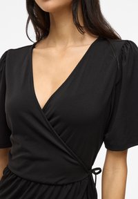 Μαύρο φόρεμα περιτυλίγματος με κοντά φουσκωτά μανίκια, που διαθέτει βαθύ V- λαιμόκοψη και δεμένη λεπτομέρεια στη μέση, φτιαγμένο από έναν απαλό, ελαφρύ ύφασμα.