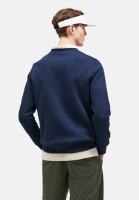 Felpa blu navy, in misto cotone, colletto tondo, con polsini e orlo a coste, indossata sopra una maglietta bianca, vista da dietro, abbinata a pantaloni verde oliva.