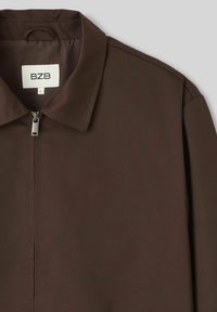 Veste zippée marron avec un design col, présentant une texture lisse et des détails de couture minimaux. L'étiquette indique "BZB" et la taille "L."