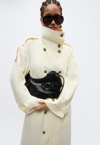 Γυναίκα με πλεξούδες στα μαλλιά φορώντας υπερμεγέθη μαύρα γυαλιά ηλίου, ένα μπεζ τραντς παλτό με ψηλό γιακά και κρατώντας μια μαύρη δερμάτινη τσάντα στο στήθος της.