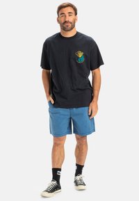 Un tricou negru din bumbac cu un imprimat grafic, asortat cu pantaloni scurți de culoare albastru deschis. Poartă adidași negri și șosete negre.