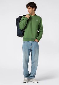 Groene trui met geribde boorden en zoom, gecombineerd met losse blauwe jeans. Het model draagt een donkerblauwe rugzak en grijze sneakers.