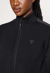 Zwarte ritsjack met hoge kraag, gestructureerde stof en een logopatch op de borst; voorzien van contrasterende grijze accenten op de schouders.