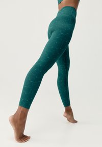 Gröna leggings med en texturerad, vågig mönstring, hög midja, figurnära design och elastiskt material. Barfota position framhävs.