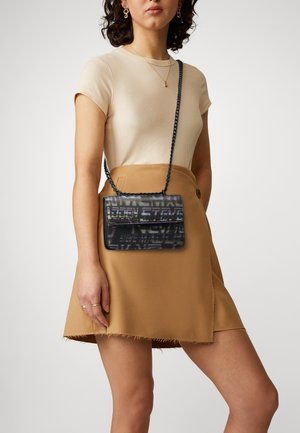 Femme aux cheveux bouclés portant un haut beige à manches courtes, une jupe portefeuille couleur sable et un sac bandoulière noir Steve Madden avec une chaîne en bandoulière.