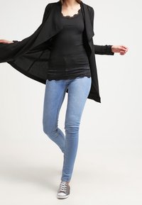 Svart öppen kardar över en svart linne med spetsdetaljer, tillsammans med ljusblå skinny jeans och grå sneakers.