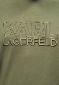 Ύφασμα ελιάς με ανάγλυφο κεντητό κείμενο "KARL LAGERFELD" και περίγραμμα κεφαλιού άνδρα με γυαλιά ηλίου και αλογοουρά μέσα στο γράμμα K.