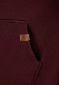 Vinrød sweatshirtstof med en ribbet tekstur, der har en frontlomme og et tanfarvet lædermærke med teksten "VIVID SENCE."