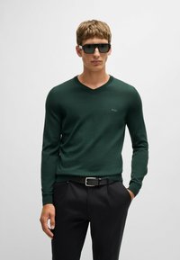 Zielony sweter w serek z miękkiego materiału, z długimi rękawami i delikatnym logiem. Noszony z czarnymi spodniami i czarnym paskiem.