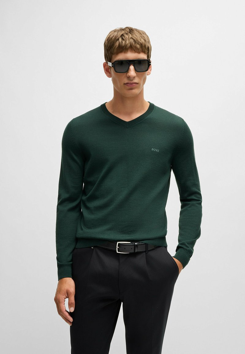 Zielony sweter w serek z miękkiego materiału, z długimi rękawami i delikatnym logiem. Noszony z czarnymi spodniami i czarnym paskiem.