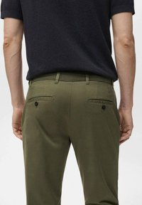 Olive green chinos with a flat front, belt loops, and two back pockets featuring button closures, paired with a black short-sleeve top.