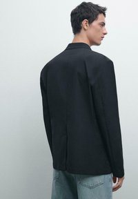 Schwarzer Blazer mit strukturiertem Design, Reverskragen und einer Rückenschlitz. Getragen über hellblauen Jeans, die eine glatte Textur aufweisen.
