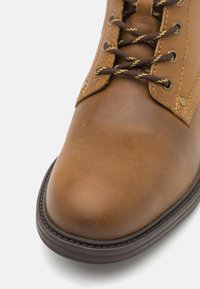 Bota de cuero marrón con una textura suave, punta redonda, costuras en contraste y suela de goma oscura. Presenta ojales de metal y cordones marrones.