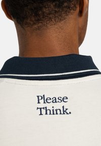Collared polo shirt in light cream with a dark navy collar, featuring navy embroidered text "Please Think." on the back. Smooth texture.