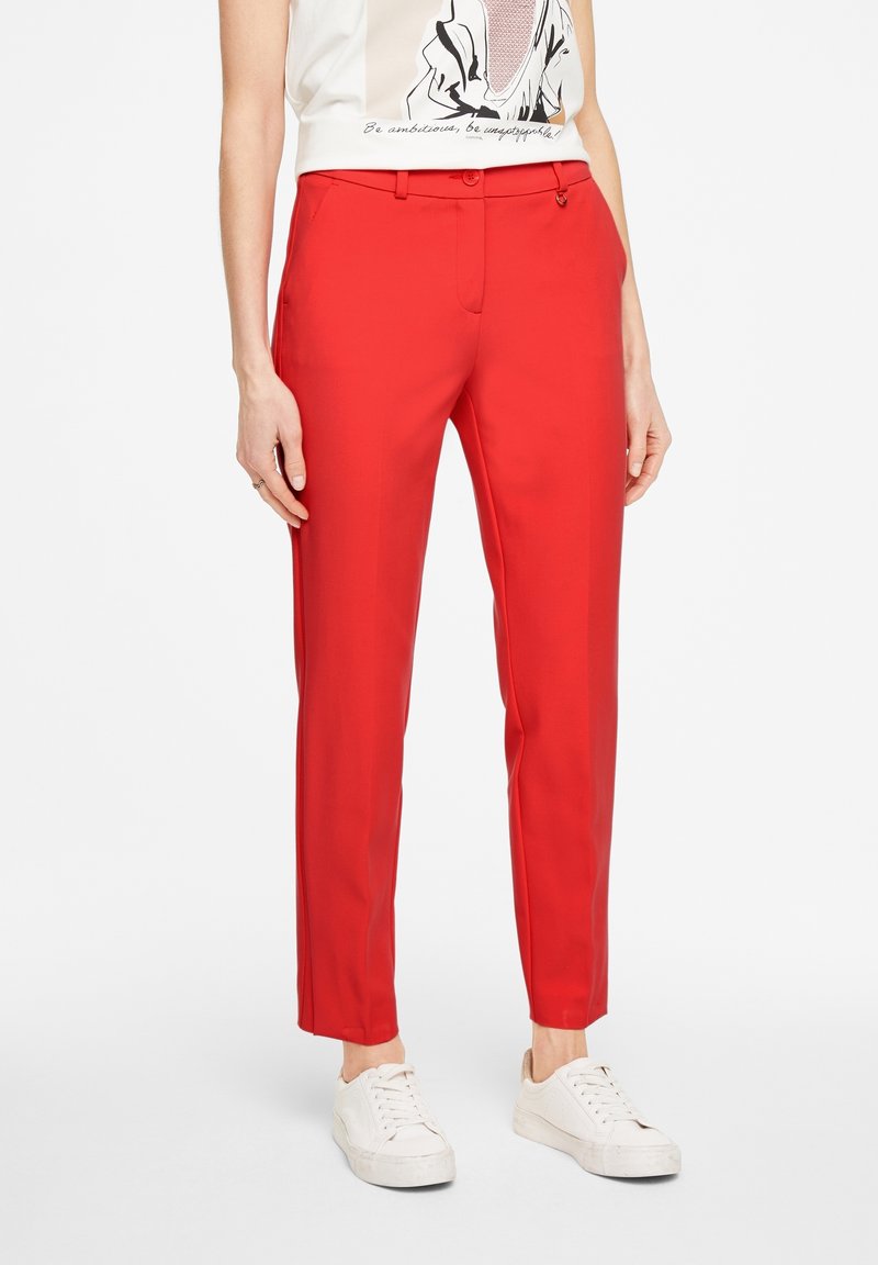 Pantalon rouge slim jusqu'à la cheville porté avec des baskets blanches et un t-shirt blanc avec un motif graphique noir et beige.