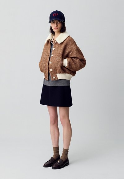 Chaqueta bomber de textura en espiga marrón con cuello de piel sintética blanca, combinada con una blusa gris, falda azul marino, calcetines marrones y mocasines negros.