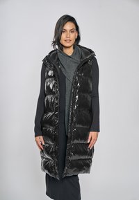 Lange schwarze Steppweste mit glänzender Textur, Reißverschluss und Kapuze. Wird über einem dunkelgrauen Oberteil getragen, mit lockerer Passform und knielanger Länge.