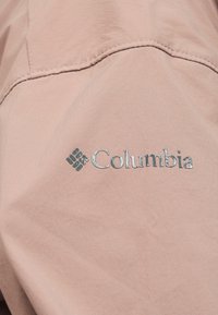 Logo Columbia en impression métallique sur tissu doux rose clair avec couture visible au-dessus du logo.