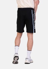 Schwarze Baumwollshorts mit geradem Schnitt. Hellblaue seitliche Streifen mit Logo-Text. Getragen mit weißen Socken und grauen Turnschuhen.