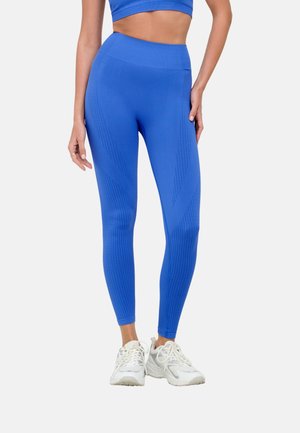 Žena v jasne modrých legínach s vysokým pásom bez švov so štruktúrovanými vzormi a bielych športových teniskách, stojaca na bielom pozadí.