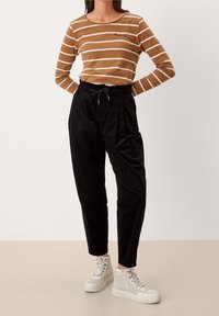 Person som har på sig en långärmad tröja med bruna och vita ränder, svarta högmidjade byxor med dragsko och vita högstövlar som står på en beige golv.