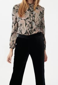 Blouse florale en tissu foncé avec col à volants et boutons, associée à un pantalon en velours noir. Manches longues et motifs paisley élaborés.