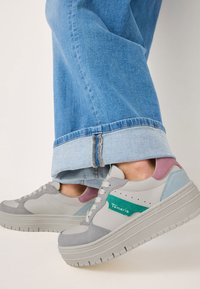 Baskets grises et pastel présentant un mélange de matériaux lisses et texturés. Semelle épaisse et superposée avec un logo vert bien visible. Jean en denim.