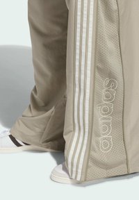 Bež trenerke s bijelim prugama sa strane, s perforiranim panelima i izvezenim detaljima brenda. Uparene s bijelim tenisicama.