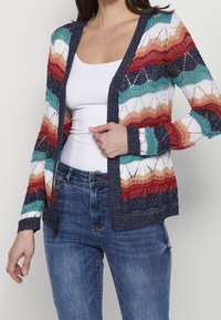Sweter kardigan w wielokolorowe paski w odcieniach czerwieni, bieli, teal i granatu, z koronkowym wzorem, otwarty z przodu oraz z długimi rękawami.