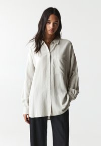 Chemise à manches longues, boutonnière de couleur gris clair, avec une poche, coupe décontractée et texture douce. Assortie à un pantalon large sombre.