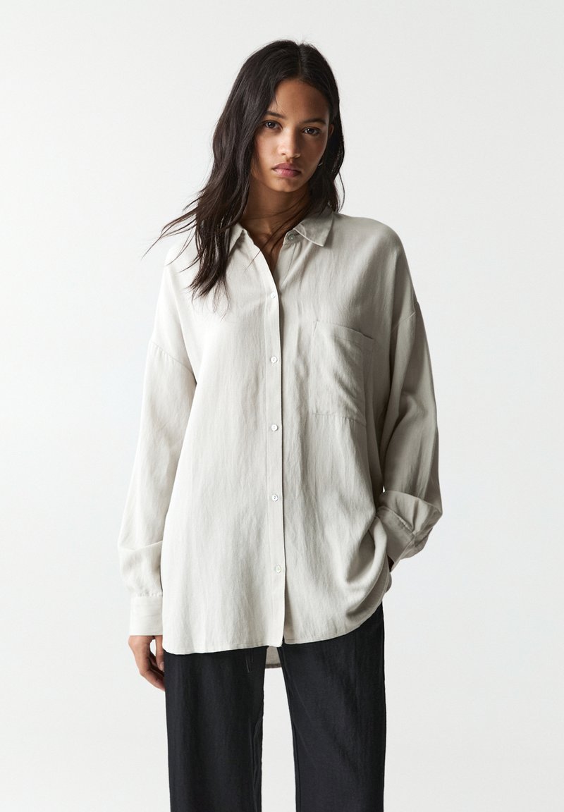 Chemise à manches longues, boutonnière de couleur gris clair, avec une poche, coupe décontractée et texture douce. Assortie à un pantalon large sombre.