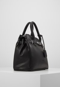 Borsa a mano in pelle nera texturizzata con doppi manici, dettaglio fibbia argentata e targhetta in pelle attaccata, posta su un piedistallo bianco su sfondo semplice.