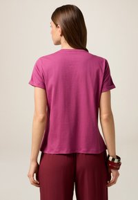 T-shirt à manches courtes et col rond en coton magenta uni. Coupe ample avec un ourlet légèrement arrondi, associé à un pantalon bordeaux ample et à des bracelets multicolores.