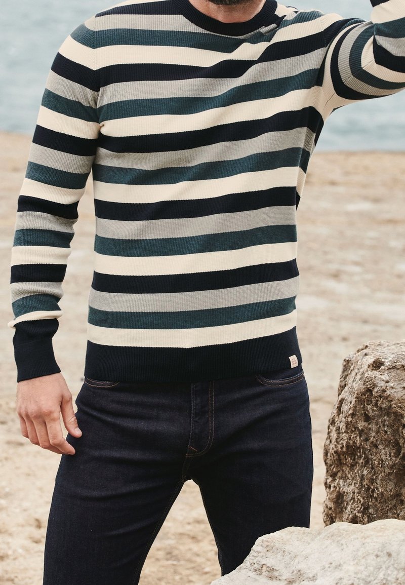 Pull en tricot avec des rayures horizontales bleu marine, vert sarcelle, gris clair et crème. Il a un col rond et des poignets côtelés. Porté avec un jean foncé.