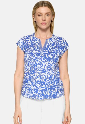 Femme portant un chemisier à manches courtes à motif floral bleu et blanc avec une petite fente en V au col, debout devant un fond uni.