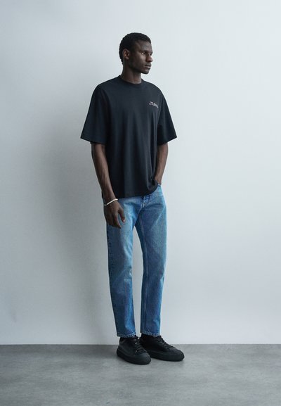 Άνδρας στέκεται μπροστά σε έναν γκρίζο τοίχο, φορώντας μια φαρδιά μαύρη κοντομάνικη μπλούζα, μπλε τζιν, μαύρα αθλητικά παπούτσια και ένα λευκό βραχιόλι με χάντρες στον αριστερό καρπό του.