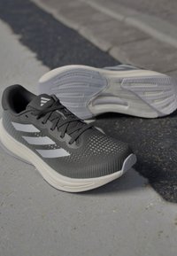 Baskets de sport grises avec un dessus en maille perforée, accents blancs et semelles rembourrées. Présente un design texturé et des lacets plats.