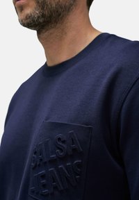 Sudadera azul marino con un bolsillo de logo en 3D texturizado. Presenta un cuello redondo y una textura de tela suave. Detalles de costura visibles en las mangas.