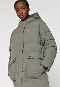 Pufinė parka striukė muted žalia spalva, su kapišonu, priekinėmis kišenėmis ir subtiliu logotipu. Minkšta tekstūra su horizontaliu siuvinėjimo dizainu.