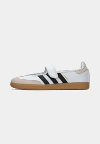 White sneaker with black stripes, a brown rubber sole, and a beige suede toe. Velcro strap across the top, minimal branding on the side.