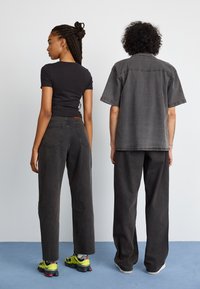 Svart tettsittende t-skjorte og løse grå denimbukser på to modeller. Det ene paret har på seg flerfargede sko, mens det andre har hvite. Enkel design, avslappet passform.