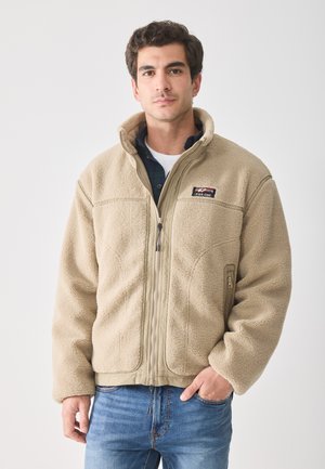 Chaqueta de forro polar beige con cierre de cremallera, cuello alto y dos bolsillos laterales con cremallera. Tejido texturizado con un diseño sencillo.