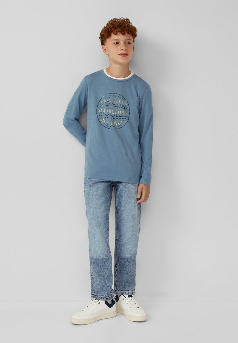 Blauwe langemouw T-shirt met een cirkelvormig "SOUND" tekstontwerp, gecombineerd met lichtblauwe jeans en witte sneakers. Soepele stof, casual pasvorm.