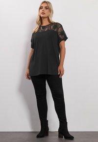 Top nero a maniche corte con sovrapposizione di pizzo sulle spalle, abbinato a pantaloni neri aderenti e stivaletti con tacco largo. Texture liscia.
